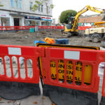A sign saying businesses open as usual obscured by a roadworks barrier.